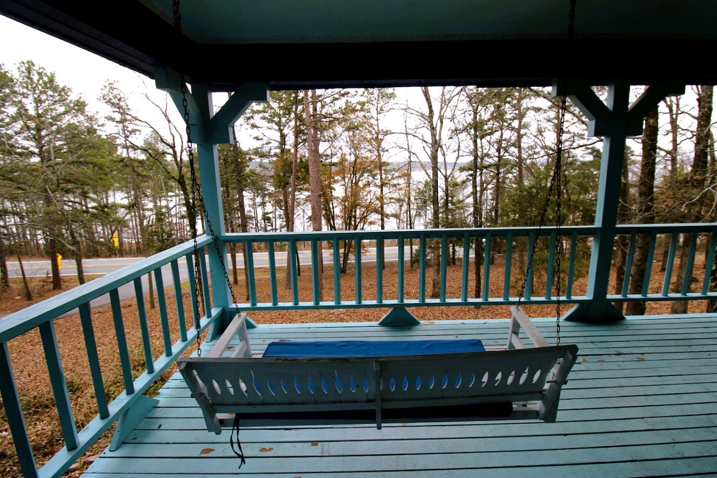 A covered porch with a bench, a railing, and a view of trees and a road.