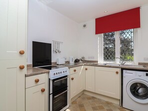 Une cuisine compacte avec des meubles blancs, une cuisinière, un four, un lave-linge et une fenêtre avec un store rouge.