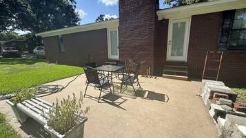A patio with a table and chairs, a concrete planter with plants, and a brick building with a door and windows.
