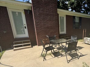 Une terrasse avec une table en verre et des chaises en métal, un bâtiment en briques avec une porte, et un petit escalier menant à l’entrée.