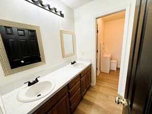 Un baño con dos lavamanos, un espejo y una puerta que da a otra habitación.
