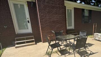 A patio with a glass table and metal chairs, a brick building with a door, and a small set of steps leading to the entrance.