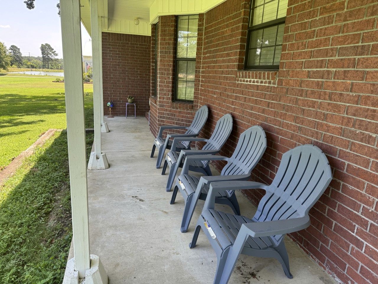 A covered outdoor seating area with rows of chairs against a brick wall, a concrete floor, and a grassy field in the background.