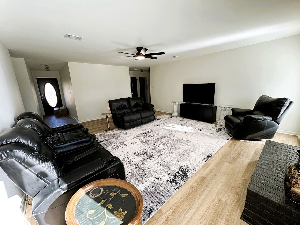 Un salon avec un canapé en cuir noir, un fauteuil noir, une table basse en bois, un tapis à motifs et un ventilateur de plafond.