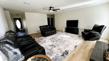 Un salon avec un canapé en cuir noir, un fauteuil noir, une table basse en bois, un tapis à motifs et un ventilateur de plafond.