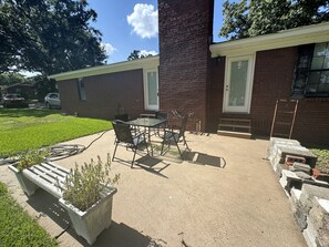 Un patio con una mesa y sillas, una jardinera de concreto con plantas y un edificio de ladrillo con una puerta y ventanas.