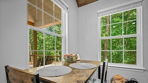 Een eetruimte met een houten tafel, twee stoelen, een mand met handdoeken en uitzicht op groen door grote ramen.
