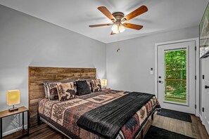 Ein Schlafzimmer mit einem hölzernen Kopfende, einer gemusterten Tagesdecke, einem Deckenventilator, Nachttischlampen und einer Tür zu einem begrünten Außenbereich.