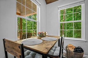 Ein Essbereich mit einem Holztisch, zwei Stühlen, einem Korb mit Handtüchern und Blick ins Grüne durch große Fenster.