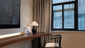 Hotellihuone, jossa on taulutelevisio, lamppu, tuoli ja ikkuna, jossa on verhot.