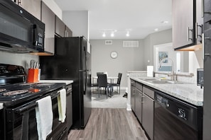A modern kitchen with dark cabinetry, a black refrigerator, a stove, and a dining area with chairs and a table. - Bright & spacious 1bedroom retreat (New Braunfels)