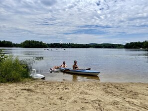 A sandy beach with a calm lake, a small boat, and two people sitting in the boat. - Spacious Cottage for a Group Getaway with Views of Harvey Lake in Northwood, New Hampshire (Northwood)