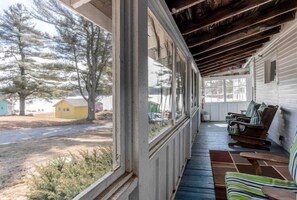 A covered porch with a view of a rural landscape, including a small yellow building and trees. - Spacious Cottage for a Group Getaway with Views of Harvey Lake in Northwood, New Hampshire (Northwood)