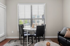 Ein Essbereich mit Tisch und Stühlen, ein Fenster mit Jalousien, eine Vase und eine Couch mit Kissen.