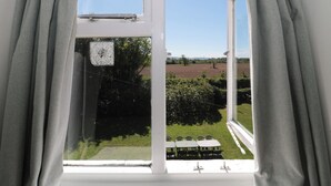 A window with curtains, a view of a green landscape, and a white building in the distance. - Walk Farmhouse (Gloucester)