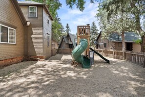 Ein Spielplatz mit Rutsche, umgeben von Holzspielgeräten und einer Schotterfläche.
