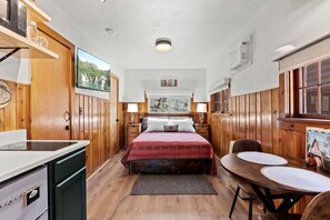 Quarto compacto com painéis de madeira, uma cama, mesinhas de cabeceira, uma pequena mesa de jantar e vista para uma floresta pela janela.