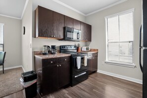 A kitchen with dark cabinets, a microwave, stove, and a window with blinds. - Cozy 2BR with modern finishes (New Braunfels)