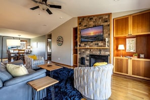 A living room with a fireplace, a television mounted on a stone wall, and a dining area in the background. - Relaxing Coastal Getaway with Bay Views in Blaine, Washington (Blaine)