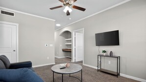 Een woonkamer met een plafondventilator, een flatscreen televisie, een ronde salontafel, een bank en een console tafel met een plant.