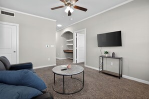 Ein Wohnzimmer mit Deckenventilator, einem Flachbildfernseher, einem runden Couchtisch, einem Sofa und einem Konsolentisch mit Pflanze.