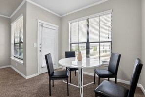 Ein Zimmer mit einem runden Tisch, vier Stühlen, einer Vase und zwei Fenstern mit Jalousien.