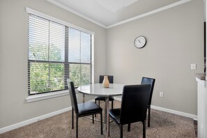 Ein Zimmer mit einem runden Tisch, vier Stühlen, einer Wanduhr und einem Fenster mit Jalousien.