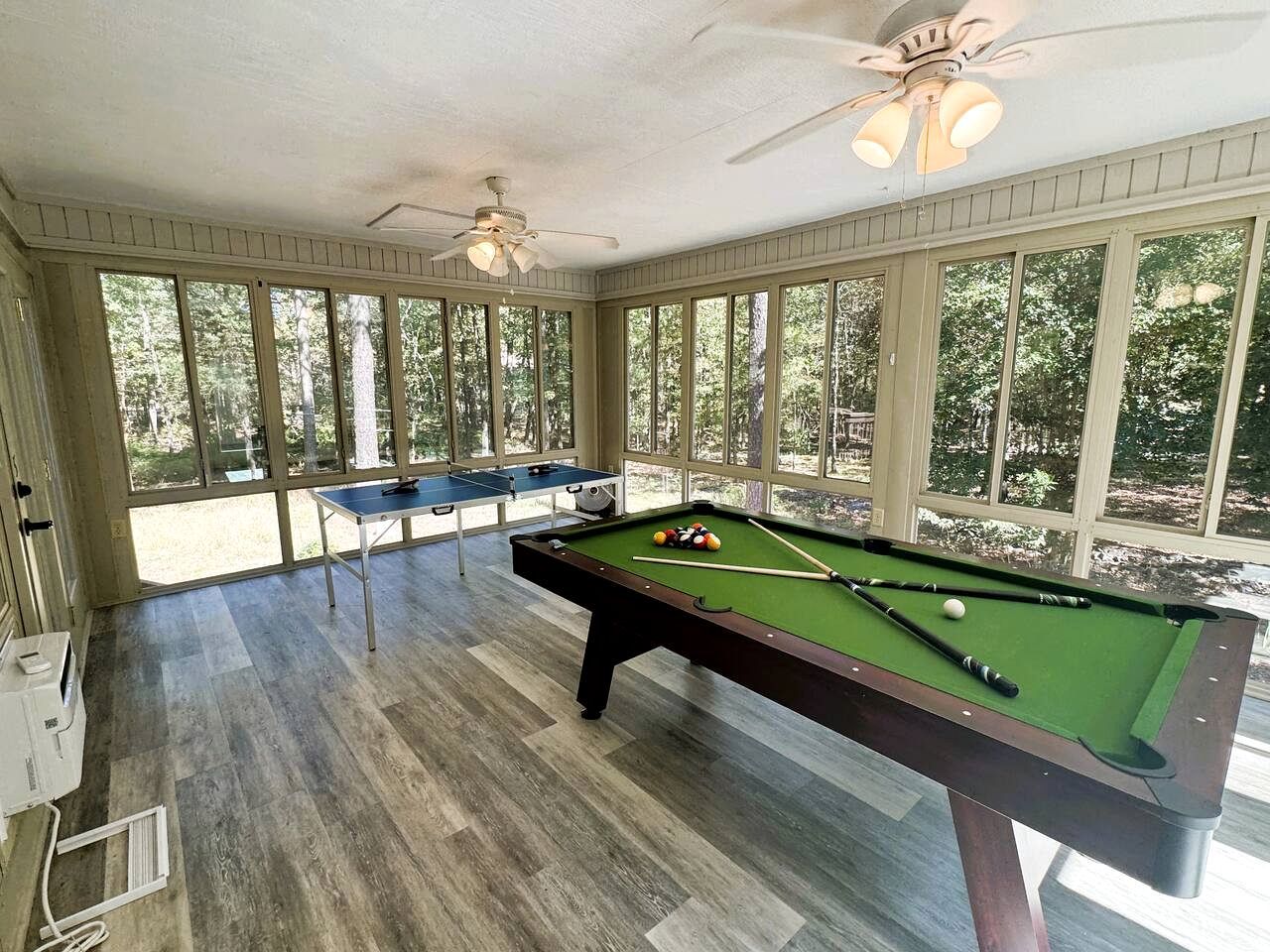 A pool table in a room with large windows, ceiling fans, and a view of trees.