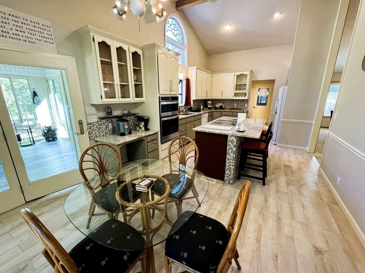 A spacious kitchen with white cabinets, a dining area with a glass table, and a view through a large window.