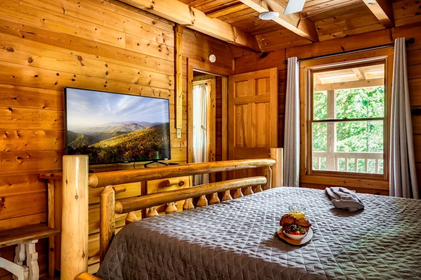 A wooden cabin room with a bed, a television mounted on the wall, and a window with a view of greenery.