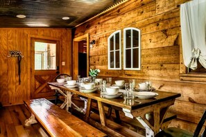 Uma sala de jantar de madeira com uma mesa comprida posta para quatro pessoas, bancos e uma janela com cortinas.
