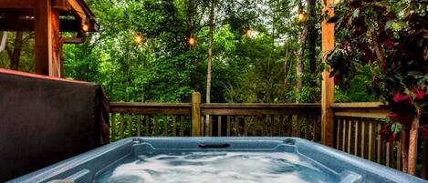 A hot tub on a wooden deck surrounded by trees, with string lights overhead.