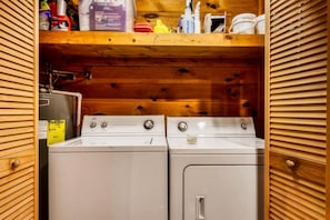 Ein Waschraum mit zwei weißen Waschmaschinen und Trocknern, Holzvertäfelung und verschiedenen Reinigungsmitteln auf Regalen.