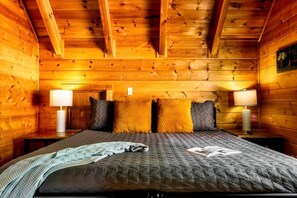 Quarto de cabana rústica de madeira, com cama, almofadas e duas luminárias de cabeceira.