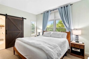 Un dormitorio con una cama grande, una mesita de noche con lámpara, una ventana con cortinas y una puerta corrediza.