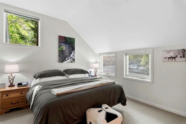 A bedroom with a bed, two bedside tables, a nightstand, a window with blinds, and a wall-mounted picture.