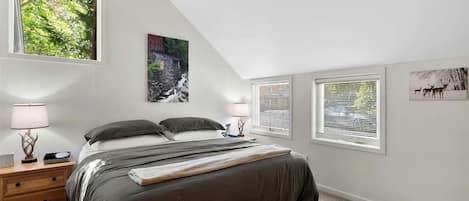 Un dormitorio con una cama, dos mesitas de noche, una mesita auxiliar, una ventana con persianas y un cuadro colgado en la pared.