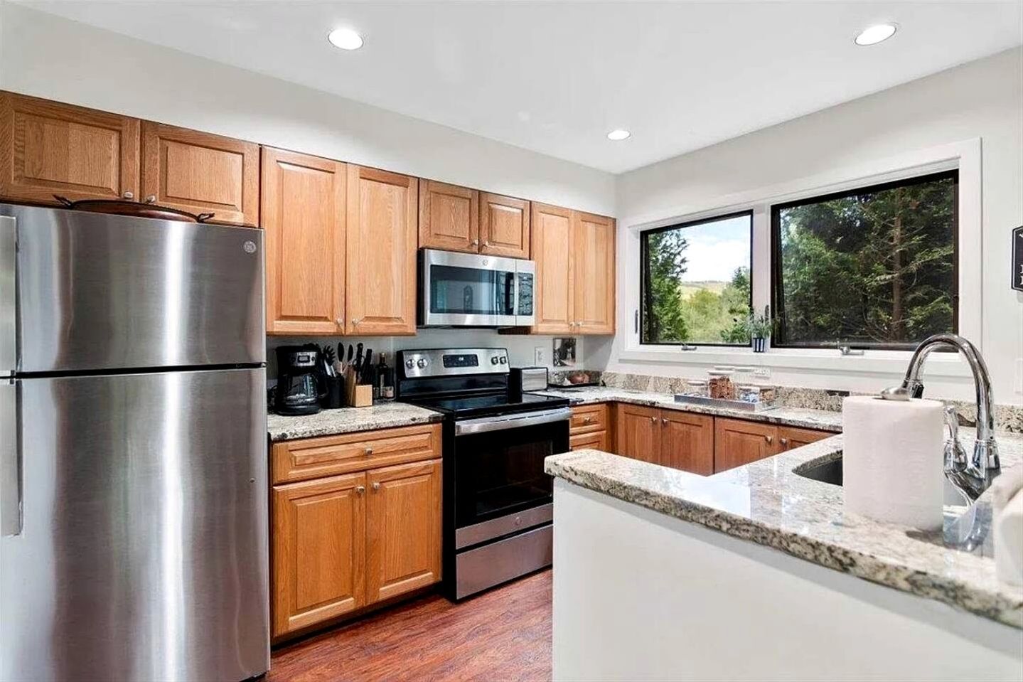 A modern kitchen with wooden cabinets, stainless steel appliances, granite countertops, and a window with a view of trees.