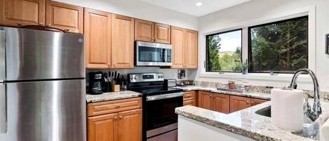 A modern kitchen with wooden cabinets, stainless steel appliances, granite countertops, and a window with a view of trees.