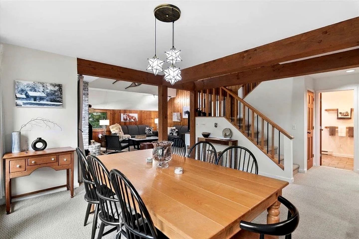 Un comedor con una mesa y sillas de madera, una escalera y una sala de estar al fondo.