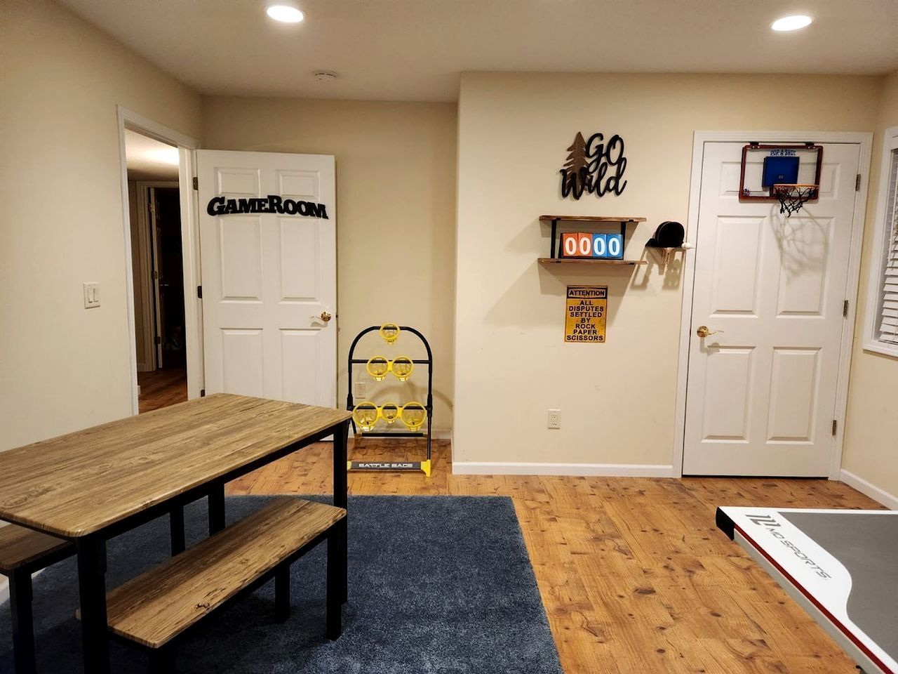 Una habitación con una mesa de madera, una alfombra azul, un estante con equipo deportivo y una puerta que indica 'GAMEROOM'.