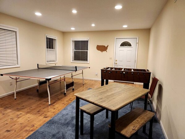 Una habitación con mesa de ping pong, mesa de futbolín y una mesa de comedor de madera con bancos.