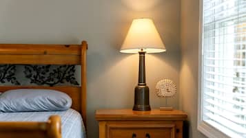 Ein Bett mit Kopfteil, ein Nachttisch mit Lampe, ein kleines Dekorationsstück und ein Fenster mit Jalousien.
