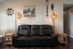 A black leather sofa with two wooden nightstands, a wall-mounted ship's wheel, a painting of mountains, and two floor lamps.