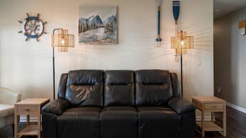 Een zwarte leren bank met twee houten nachtkastjes, een aan de muur bevestigd schipwiel, een schilderij van bergen en twee vloerlampen.