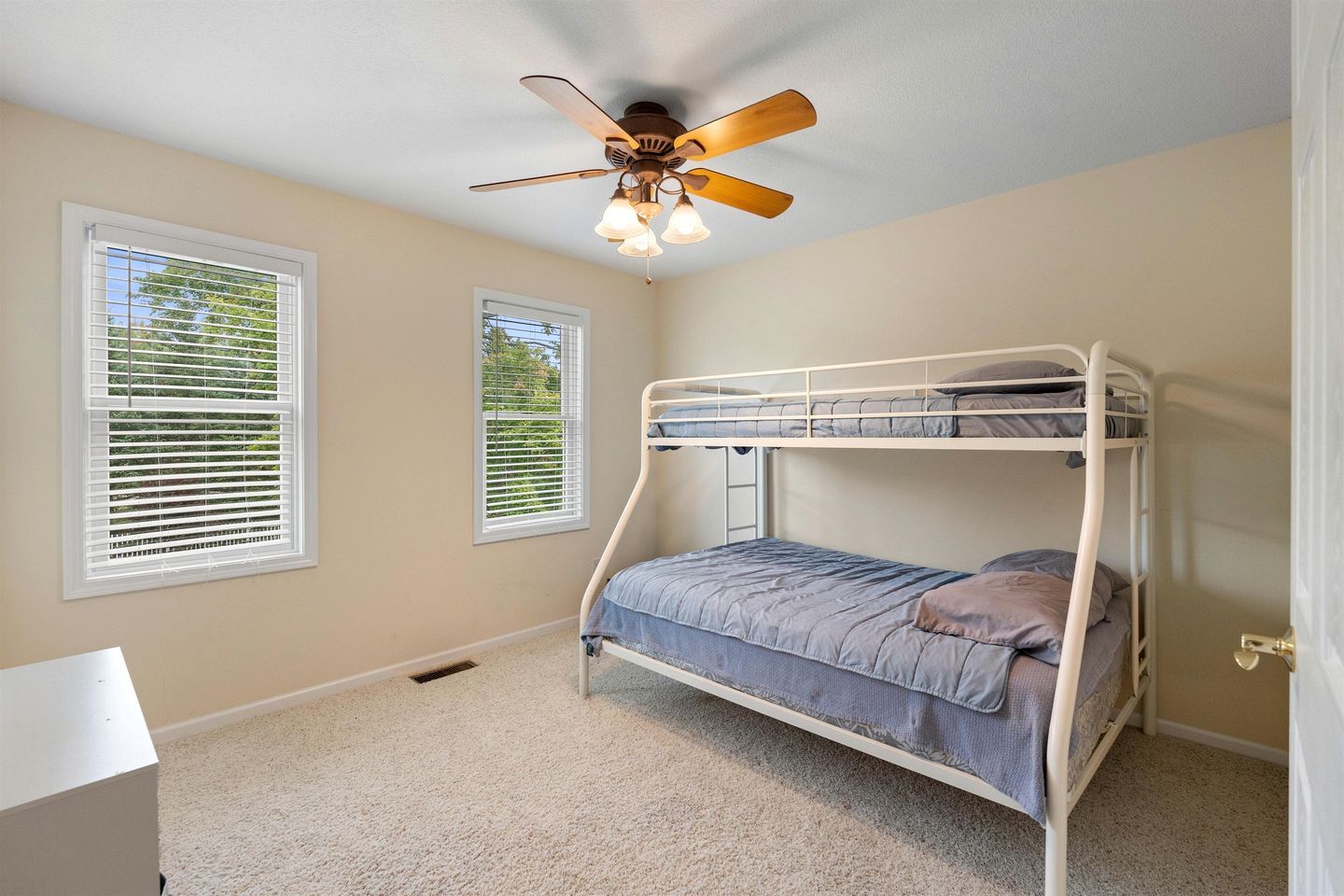 A bunk bed room with a ceiling fan, two windows with blinds, and a carpeted floor.