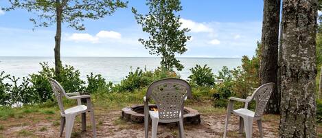A beachside seating area with three chairs and a fire pit, surrounded by trees and a view of the ocean.