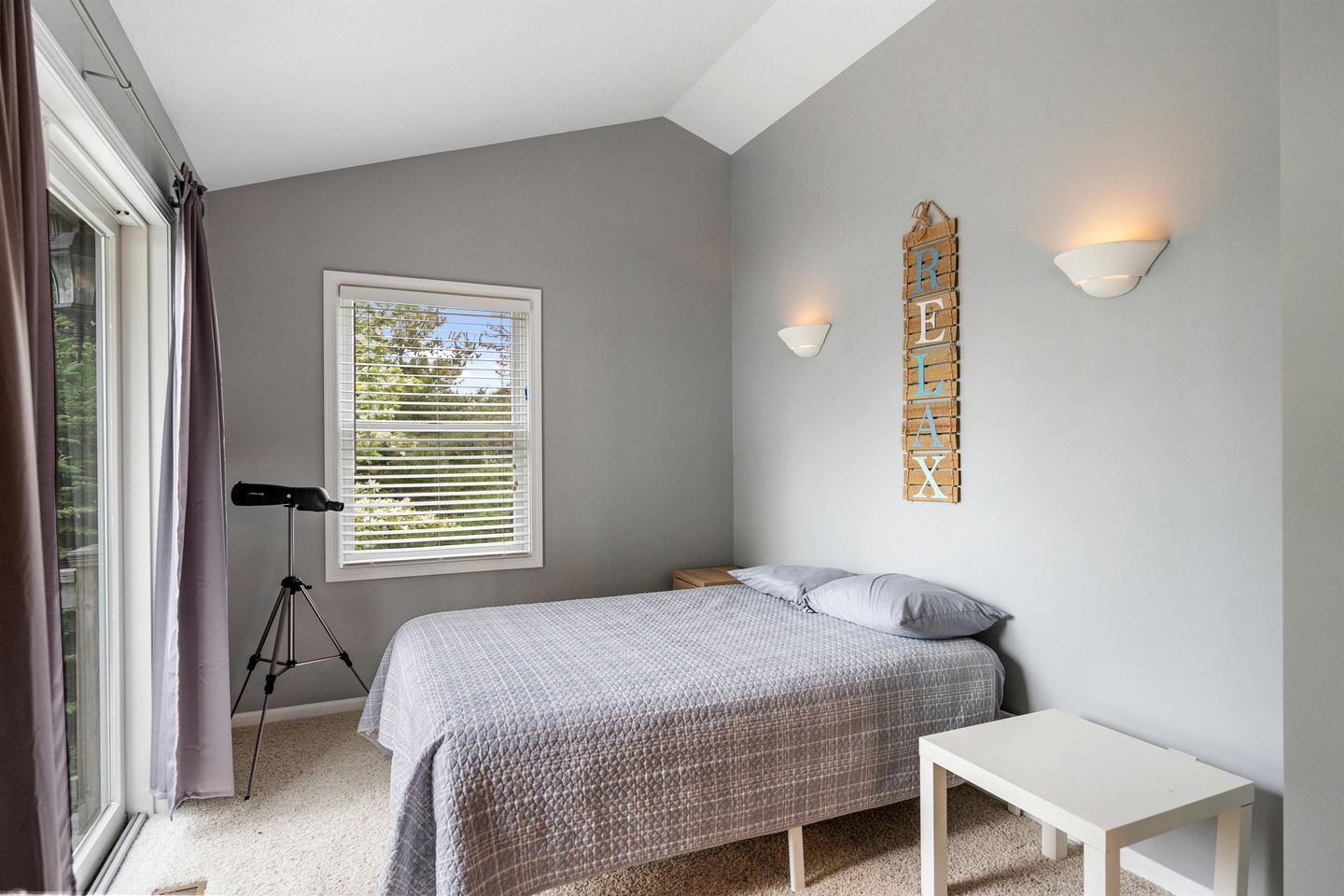 A bedroom with a bed, a small bedside table, a window with blinds, a tripod, and a wall-mounted light fixture.