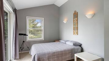 A bedroom with a bed, a small bedside table, a window with blinds, a tripod, and a wall-mounted light fixture.