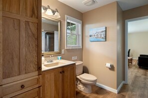 Un baño con tocador de madera, lavamanos, inodoro y ventana con persianas.